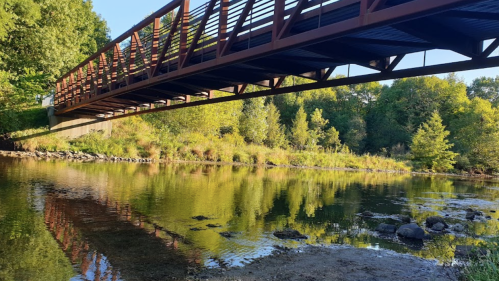 A metal bridge spans a calm river, reflecting greenery and trees on the water's surface.