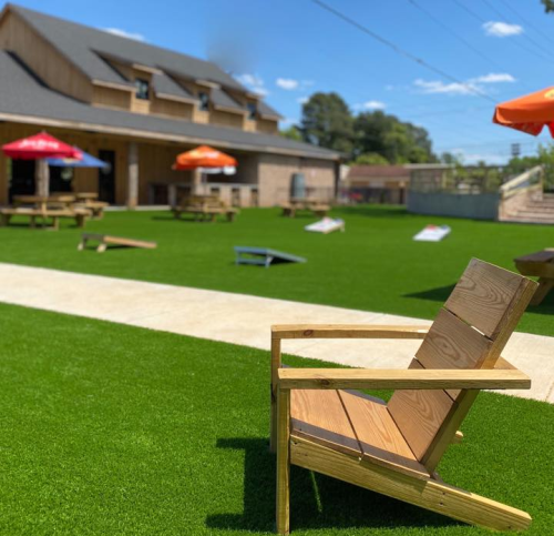 A wooden chair on green artificial grass, with picnic tables and umbrellas in the background of a sunny outdoor space.