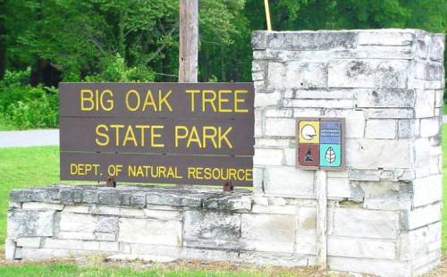 Sign for Big Oak Tree State Park, featuring stone structure and natural resource department emblem. Green surroundings.