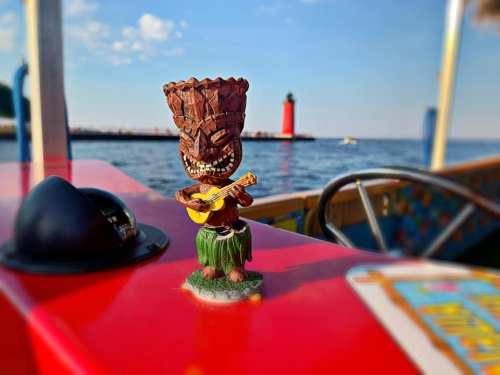 A tiki figurine playing a guitar on a red table, with a lighthouse and water in the background.