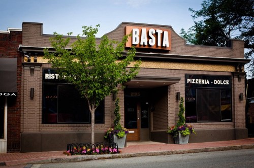 Exterior of Basta restaurant featuring a brick facade, large sign, and flower planters on a street.