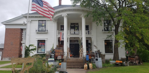 A historic white house with columns, an American flag, and various items displayed outside, including a sign that says "Open."