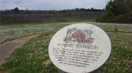 A sign titled "Winter Warmth" in a grassy area with trees and a distant landscape under a cloudy sky.