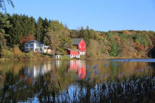 A serene lake scene with a red house and a white house surrounded by trees and autumn foliage, reflecting in the water.