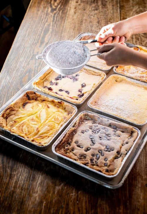 A person sifts powdered sugar over six trays of baked desserts on a wooden table.
