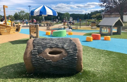 A colorful playground with a log-shaped structure, grassy areas, and various play equipment under a blue sky.