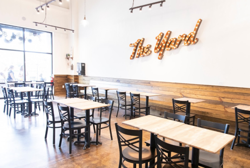 A modern restaurant interior with wooden accents, empty tables, and a lit sign reading "The Yard" on the wall.