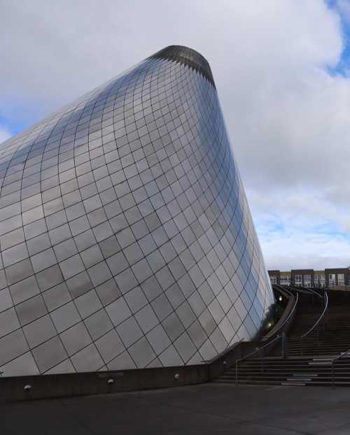 A modern, curved building with a reflective surface, set against a cloudy sky. Steps lead up to its entrance.