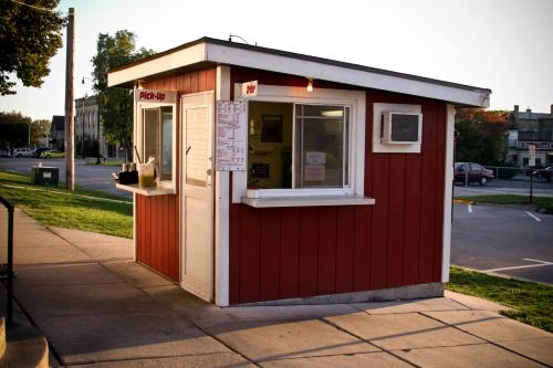 A small red food stand with a pickup window, located on a sidewalk near a grassy area and parking lot.