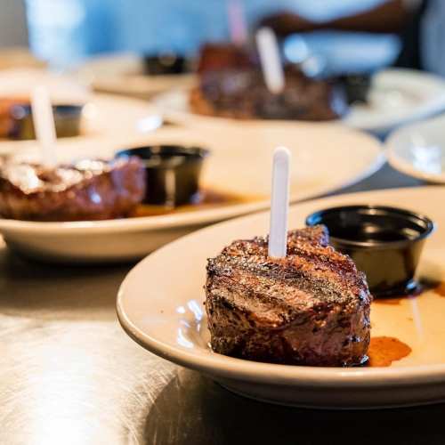 Plates of grilled steak with a dipping sauce, garnished with a white pick, on a restaurant countertop.