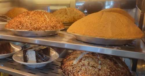 Two large, round cakes with toasted coconut topping on a display shelf, alongside a slice of chocolate cake.