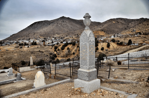 A gray tombstone stands in a cemetery with a mountainous landscape and a small town in the background under a cloudy sky.