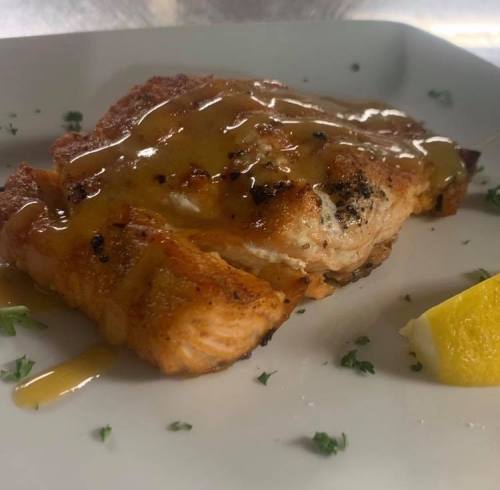 A piece of grilled salmon drizzled with a golden sauce, garnished with parsley and a wedge of lemon on a white plate.