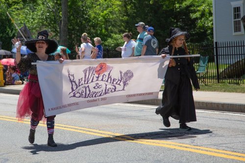 Two women in costumes march in a parade, holding a banner for a festival titled "Bewitched & Bedazzled."