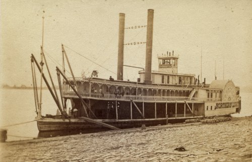 A historic steamboat docked by the river, featuring two tall smokestacks and intricate wooden details.