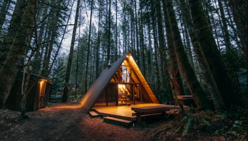 A cozy A-frame cabin nestled among tall trees in a serene forest setting, illuminated by warm lights.