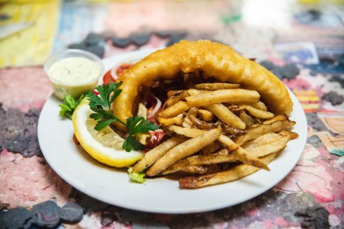 A plate of golden fried fish, crispy fries, a lemon wedge, and a small cup of dipping sauce on a colorful table.