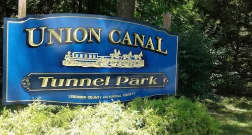 Sign for Union Canal Tunnel Park, featuring blue background and gold lettering, surrounded by greenery.