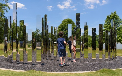 Two children play among tall, reflective pillars in a sunny park, surrounded by greenery and blue skies.