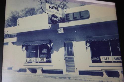Black and white photo of a vintage service station with a café, featuring signs for gas, oil, and Coca-Cola.