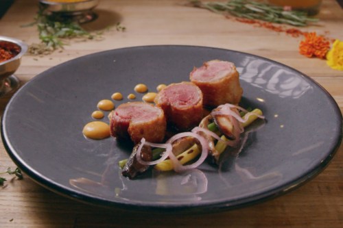 Three pieces of meat wrapped in crispy coating, served with sauce and garnished with vegetables on a dark plate.