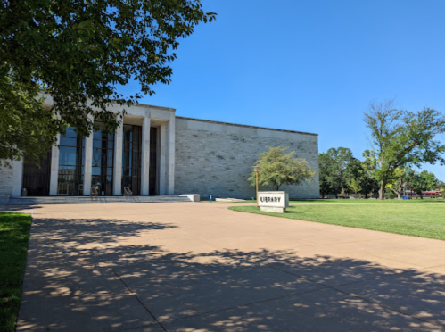 A modern library building with large windows, surrounded by green grass and trees under a clear blue sky.