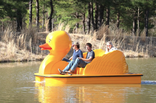 Three people enjoy a ride on a large yellow duck-shaped paddle boat on a calm lake surrounded by trees.