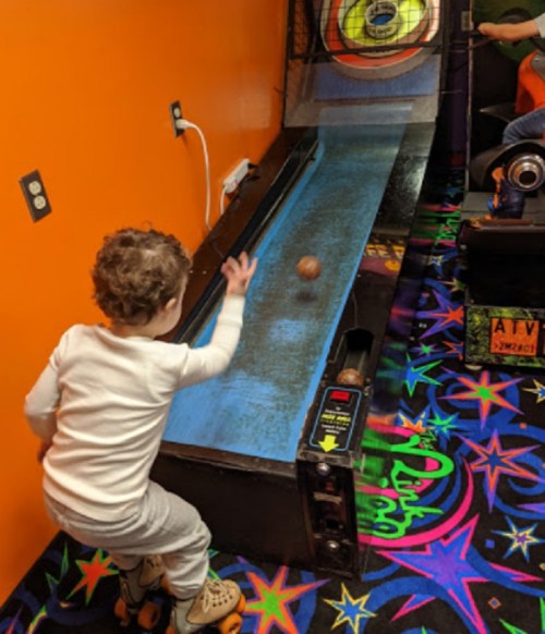 A child in pajamas rolls a ball down a blue arcade game lane with colorful carpet in the background.