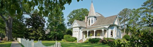 A charming Victorian-style house with a pointed turret, surrounded by greenery and a white picket fence.