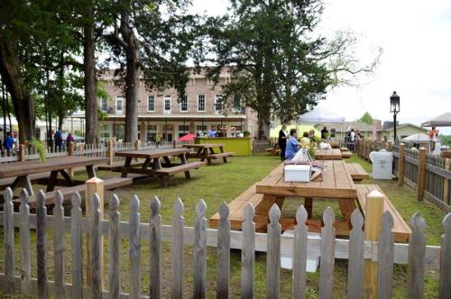 Outdoor seating area with wooden picnic tables, surrounded by a picket fence, and a building in the background.
