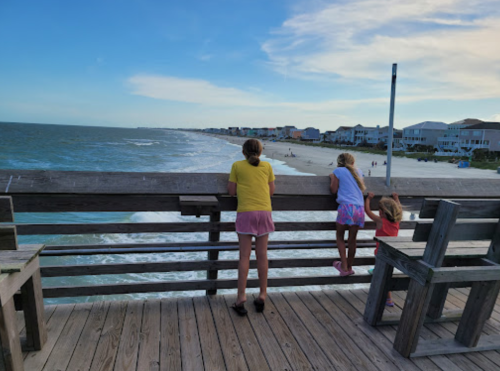 Three girls stand on a wooden pier, looking out at the ocean and beach under a blue sky with scattered clouds.
