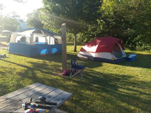 Two tents set up on a grassy campsite, with a cooler and fishing gear nearby, surrounded by trees and sunlight.