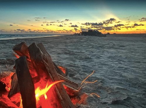 A beach bonfire glows in the foreground with a truck parked nearby, set against a vibrant sunset sky.