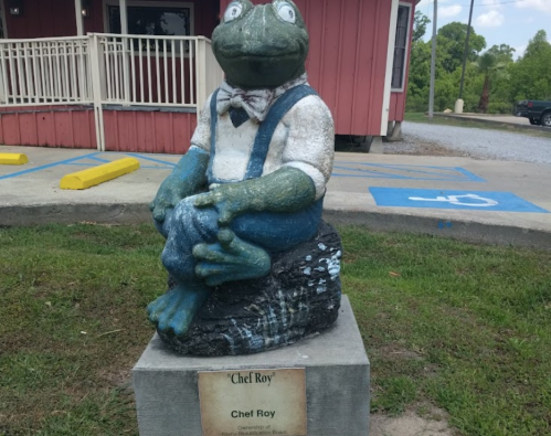 A frog statue wearing a bow tie and suspenders, sitting on a rock, with a plaque reading "Chef Roy."