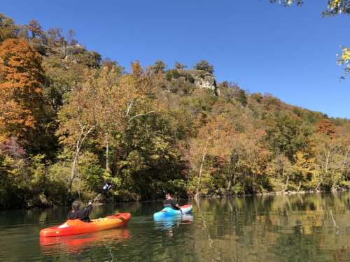 Two kayakers paddle on a calm river surrounded by vibrant autumn foliage and rocky cliffs.
