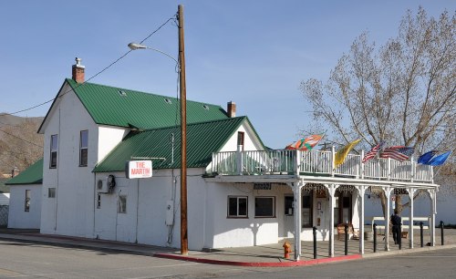 A white building with a green roof, featuring a porch and flags, located on a street with a clear blue sky.