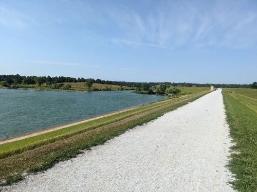 A gravel path alongside a calm lake, with green fields and trees in the background under a clear blue sky.