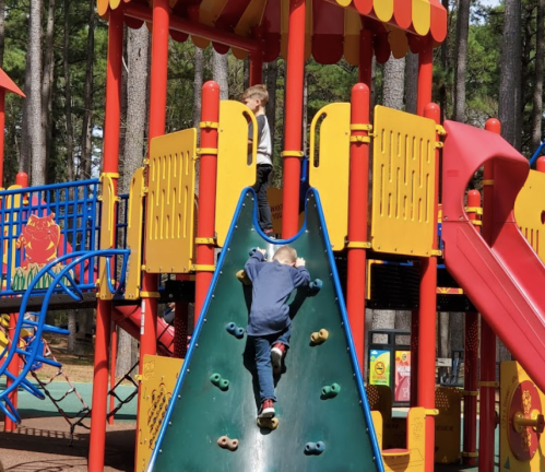 Two children play on a colorful playground structure, one climbing a slide while the other stands at the top.