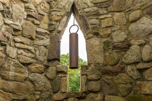 A metal bell hangs in a stone archway, surrounded by textured stone walls and greenery visible outside.