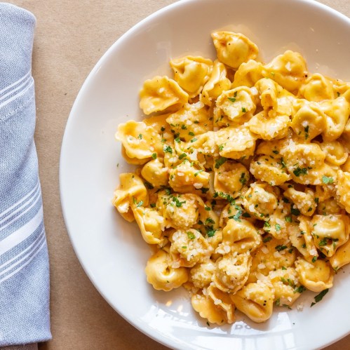 A bowl of creamy pasta with small, folded shapes, garnished with parsley and grated cheese, on a beige surface.