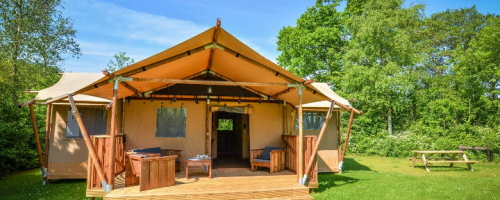 A spacious glamping tent with wooden decks, surrounded by greenery and a picnic table nearby under a clear blue sky.