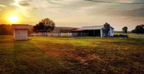 A serene farm scene at sunset, featuring a barn, silo, and a small shed on lush green grass.