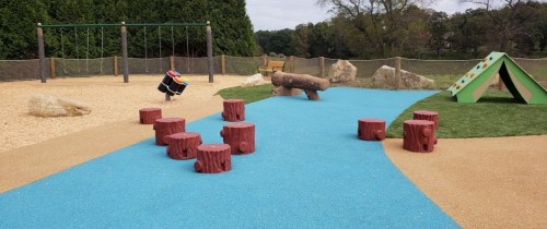 A colorful playground with log stepping stones, a climbing structure, and swings in a grassy area.