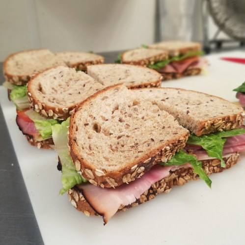 A close-up of sliced sandwiches with whole grain bread, lettuce, tomato, and ham, arranged on a white cutting board.
