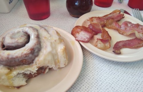 A cinnamon roll with icing on a plate next to a plate of crispy bacon strips.