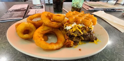 A plate featuring a stacked burger with cheese and toppings, accompanied by onion rings and waffle fries.