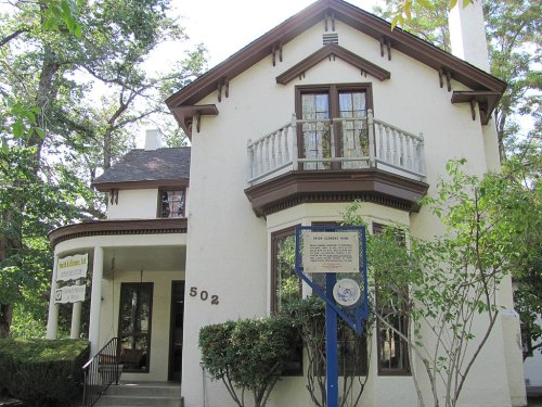 Historic two-story house with a balcony, surrounded by trees, featuring a sign and a welcoming entrance.