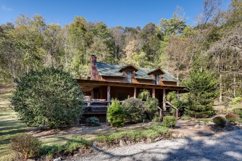 A charming log cabin surrounded by lush greenery and trees, featuring a spacious porch and a gravel pathway.