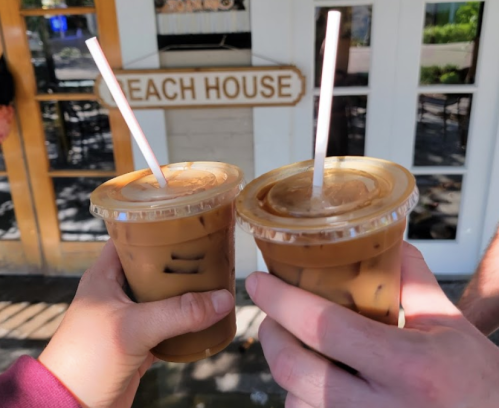 Two hands holding iced coffee cups with straws in front of a sign that reads "Beach House."