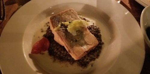 A plate of salmon on a bed of lentils, garnished with leeks and a tomato, served in a dimly lit setting.